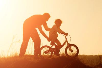 Adult helping child ride bike, sunset.