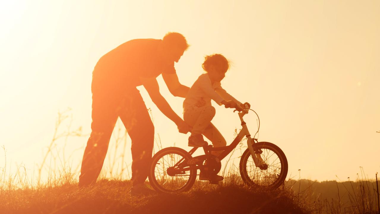 Adult helping child ride bike, sunset.
