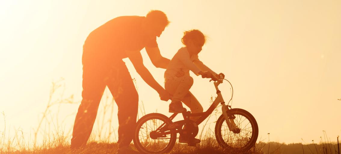 Adult helping child ride bike, sunset.