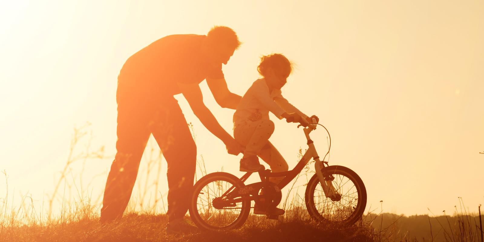 Adult helping child ride bike, sunset.